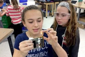 AJ-makerpsace-KJ-CC2 | MiddleWeb Two girls in a classroom examine a small electronic circuit board; one holds the device while the other watches, wearing safety goggles.