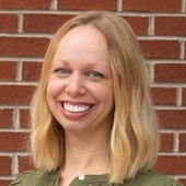 Halley-Bowman | MiddleWeb Portrait of a smiling woman with shoulder-length blonde hair wearing a green blouse, standing in front of a brick wall.